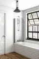 Badewanne mit verchromter Armatur, Duschkopf und Handbrause. Wände mit weißen, rechteckigen Fliesen gefliest. Fenster mit schwarzem Rahmen, Holzboden in warmer Farbe. Produkt von Ideal Standard.